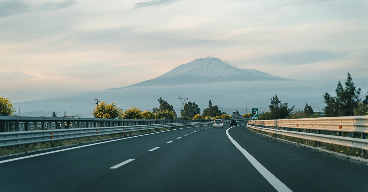 Tratto autostradale con vegetazione fiorita lungo il guardrail e un grande vulcano innevato sullo sfondo